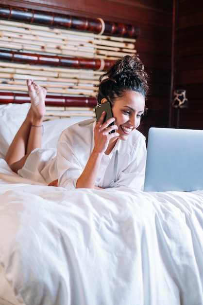 Mujer bonita con teléfono y computadora portátil en la cama