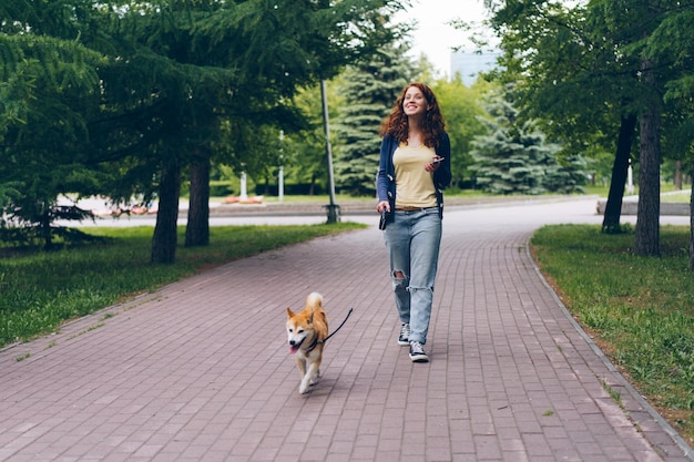 Mujer bonita con teléfono inteligente caminando perro shiba inu en el parque el día de verano sonriendo