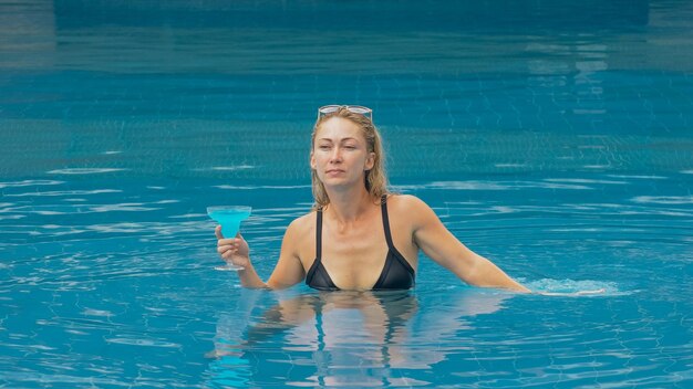 La mujer bonita de tiro medio ancho bebiendo licor de alcohol de cóctel azul en la piscina del hotel Retrato de chica sexy en la piscina al aire libre