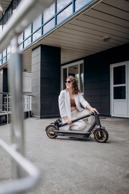 Mujer bonita con traje blanco doblando su scooter eléctrico después de un paseo mientras está de pie en el fondo de un edificio moderno