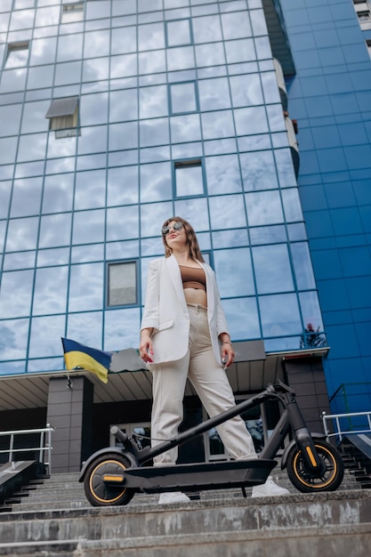 Mujer bonita con traje blanco de pie con su scooter eléctrico doblado después de un paseo en el fondo de un edificio moderno