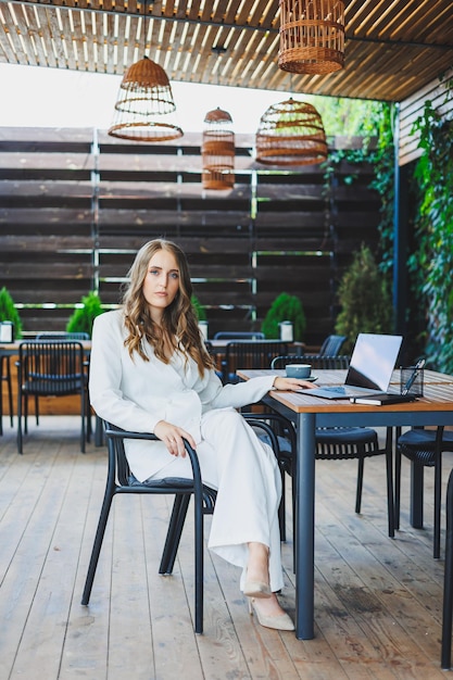 Una mujer bonita con un traje clásico blanco trabaja con una computadora portátil en una terraza abierta en una cafetería Una mujer con ropa de negocios elegante trabaja de forma remota en una computadora portátil en una cafetería de verano