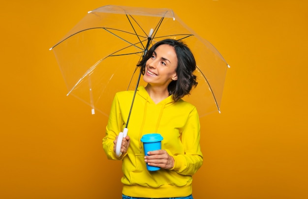 mujer bonita vestida con una sudadera con capucha amarilla, bajo un paraguas transparente, sosteniendo una taza azul en su mano izquierda, interesada en algo.