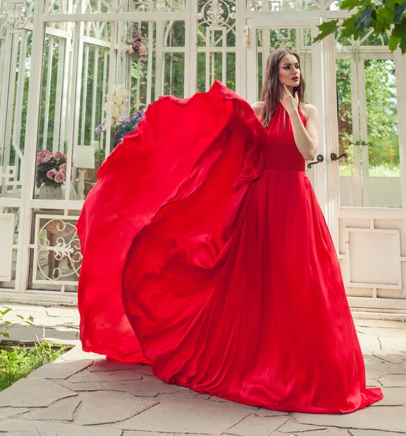 Foto mujer bonita en vestido de seda rojo de pie cerca del pabellón de verano blanco