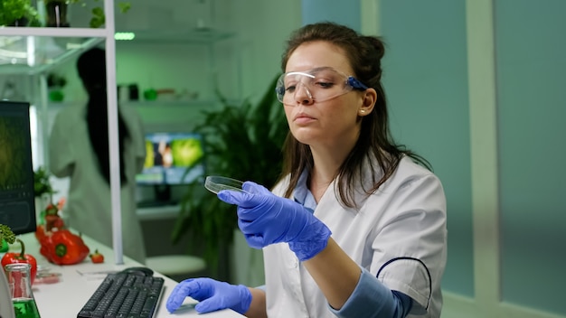 Mujer botánica mirando placa de Petri con muestra de hoja comprobando la prueba gmo escribiendo investigador científico