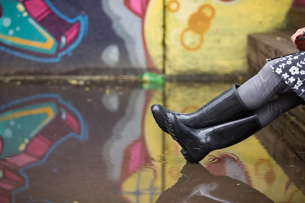 Mujer con botas de goma grises sentada en el banco después de la lluvia Par de botas de goma grises en un charco grande