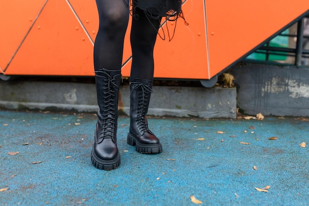 Una mujer con botas negras se para frente a una pared naranja.