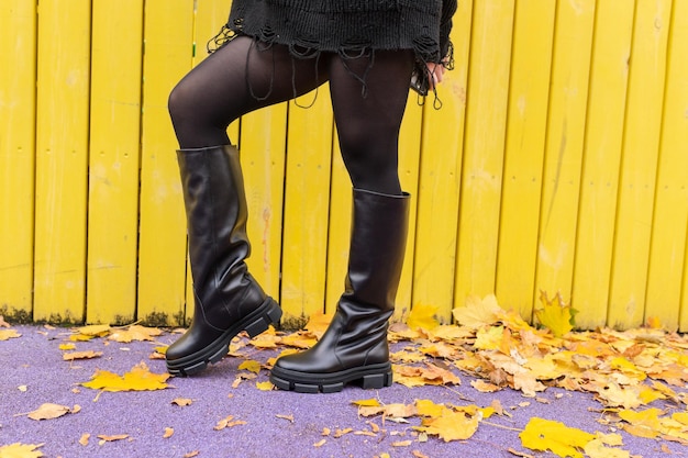 Una mujer con botas negras se para frente a una valla de madera amarilla.