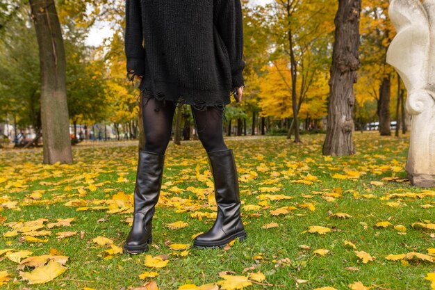 Una mujer con botas negras se para en la hierba frente a un árbol con hojas amarillas.