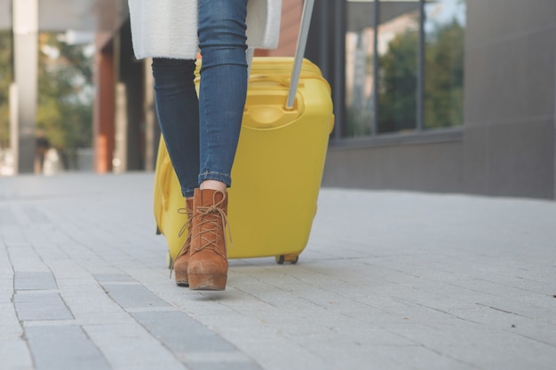 Mujer con botines en un sombrero con una maleta amarilla con un abrigo beige caminando cerca del hotel.