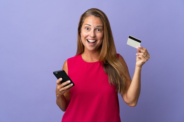 Mujer brasileña de mediana edad aislada en la pared púrpura comprando con el móvil y sosteniendo una tarjeta de crédito con expresión de sorpresa