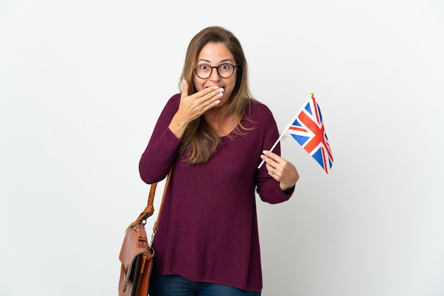 Mujer brasileña de mediana edad sosteniendo una bandera del Reino Unido aislada en la pared blanca feliz y sonriente cubriendo la boca con la mano