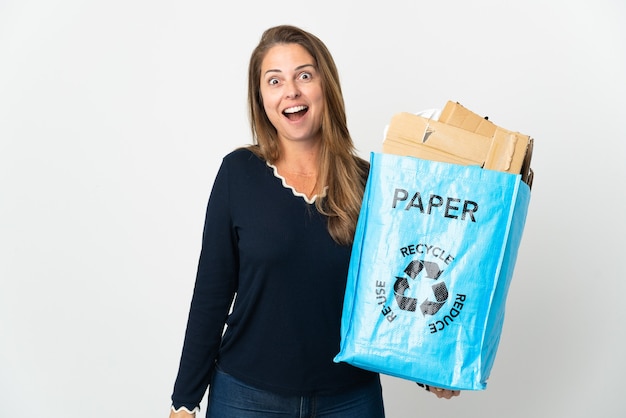 Mujer brasileña de mediana edad sosteniendo una bolsa de reciclaje llena de papel para reciclar sobre una pared aislada con expresión facial sorpresa