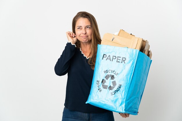 Mujer brasileña de mediana edad sosteniendo una bolsa de reciclaje llena de papel para reciclar sobre una pared aislada frustrada y cubriendo las orejas