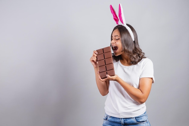 Mujer brasileña con orejas de conejo de Pascua sonriendo alegremente sosteniendo una barra de chocolate de Pascua