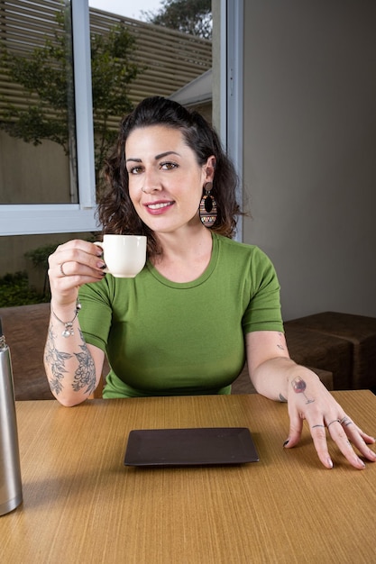 Mujer brasileña sonriente y tatuada sentada y sosteniendo una taza de café