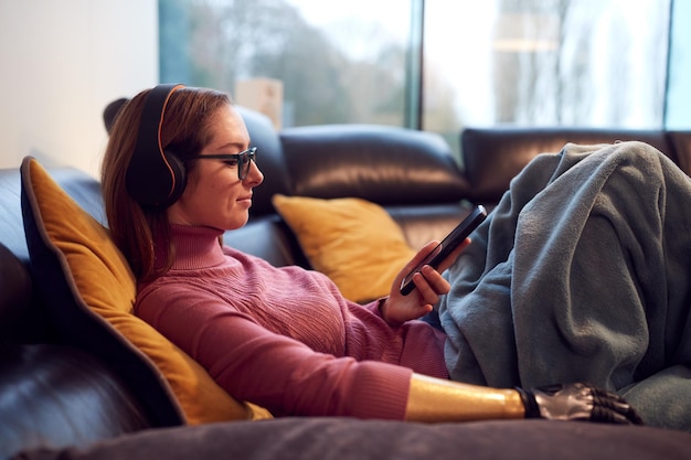 Mujer con brazo protésico usando auriculares inalámbricos escuchando música en el teléfono móvil en el sofá