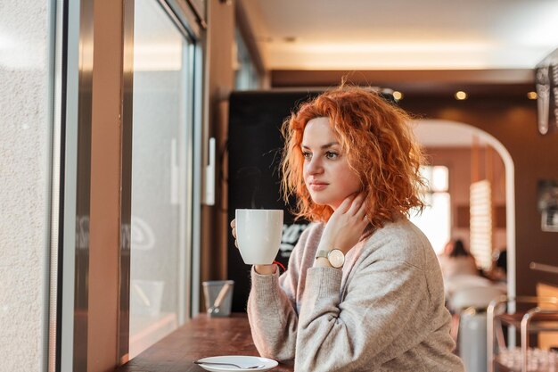 Mujer brillante joven rizada pelirroja con ojos verdes bebe café caliente en un café acogedor Hermosa modelo en ropa elegante Europa