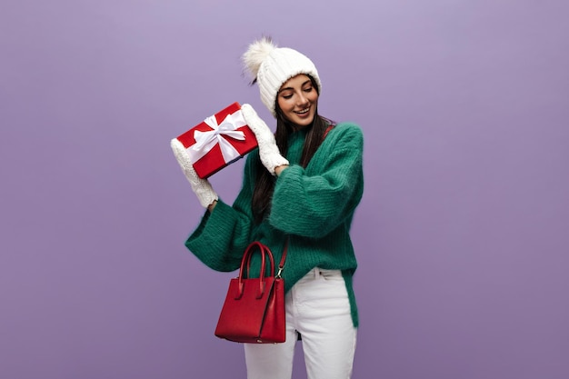 Mujer bronceada con suéter verde y pantalón blanco sacude la caja de regalo roja y sonríe Chica feliz con sombrero y guantes posa sobre fondo púrpura