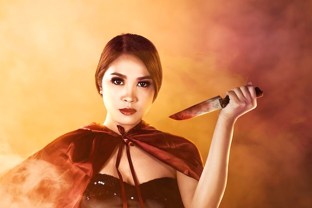 Mujer bruja asiática con un manto sosteniendo un cuchillo de pie con un fondo dramático