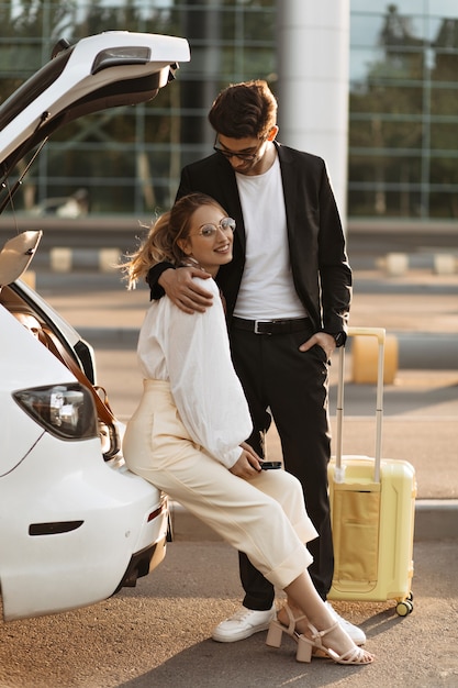 Foto mujer de buen humor en blusa blanca y pantalón beige sonríe y se sienta en el coche cerca del aeropuerto