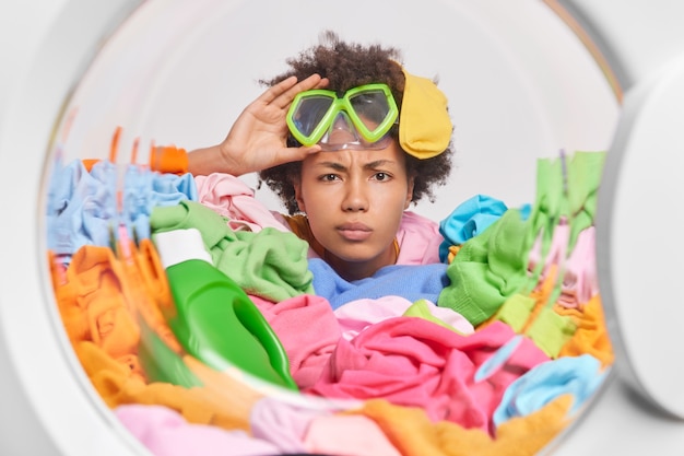 Mujer con cabello afro lleva poses de máscara de snorkel a través de la puerta de la lavadora rodeada de ropa multicolor y detergente lava la ropa en casa