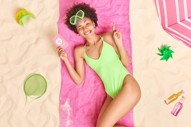 Foto mujer con cabello afro tiene pasaporte sueña con vacaciones en el extranjero mantiene los dedos cruzados viste traje de baño pasa tiempo libre en la playa con diferentes artículos alrededor. vacaciones de verano