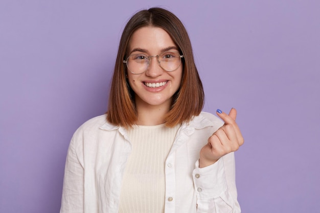 Una mujer de cabello bastante oscuro con una sonrisa suave hace que el coreano exprese su amor por ti usando una camisa blanca posando aislada sobre un fondo púrpura Gesto de minicorazón