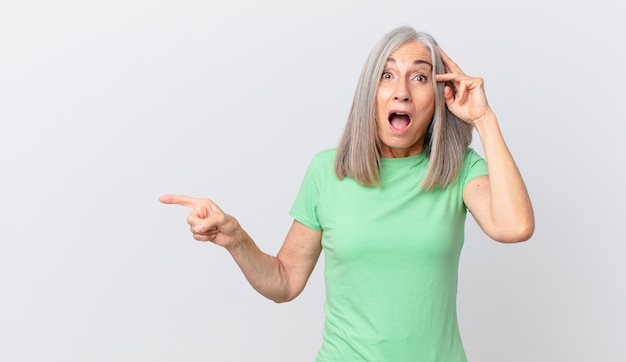 Mujer de cabello blanco de mediana edad que parece feliz, asombrada y sorprendida y apuntando hacia un lado