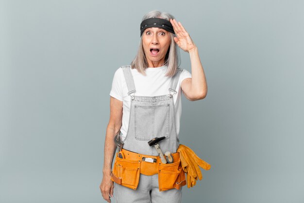 Mujer de cabello blanco de mediana edad que parece feliz, asombrada y sorprendida y con ropa de trabajo y herramientas. concepto de limpieza