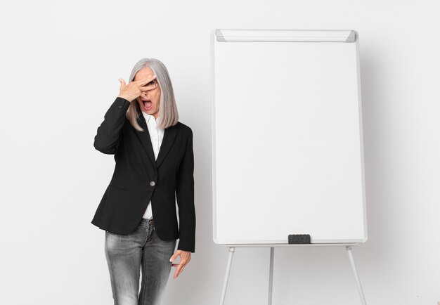 Mujer de cabello blanco de mediana edad que parece sorprendida, asustada o aterrorizada, cubriendo la cara con la mano y un espacio de copia de tablero. concepto de negocio