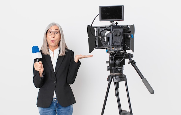 Mujer de cabello blanco de mediana edad que parece sorprendida y conmocionada, con la mandíbula caída sosteniendo un objeto y sosteniendo un micrófono. concepto de presentador de televisión