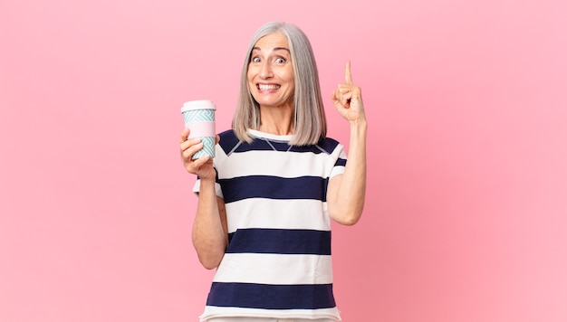 Mujer de cabello blanco de mediana edad que se siente como un genio feliz y emocionado después de darse cuenta de una idea y sostener un recipiente de café para llevar