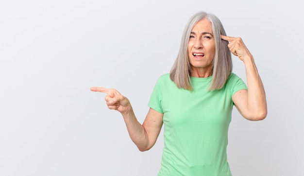 Mujer de cabello blanco de mediana edad que se siente confundida y perpleja, mostrando que estás loco y apuntando hacia un lado