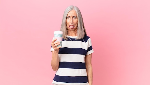 mujer de cabello blanco de mediana edad que se siente disgustada e irritada y saca la lengua y sostiene un recipiente de café para llevar
