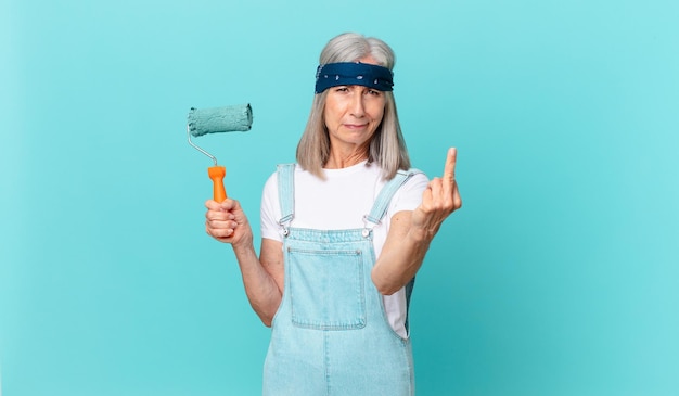 Mujer de cabello blanco de mediana edad que se siente enojada, rebelde y agresiva con un rodillo pintando una pared