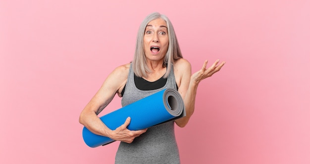Mujer de cabello blanco de mediana edad que se siente extremadamente conmocionada y sorprendida y sosteniendo una estera de yoga. concepto de fitness