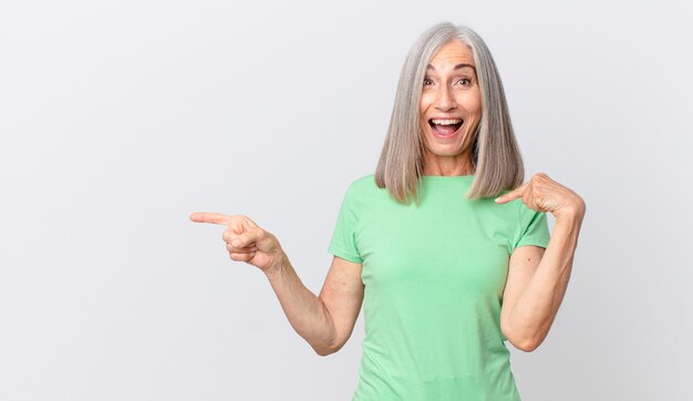 Mujer de cabello blanco de mediana edad que se siente feliz y apuntando a sí misma con un emocionado y apuntando hacia un lado