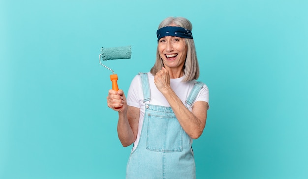 Mujer de cabello blanco de mediana edad que se siente feliz y enfrenta un desafío o celebra con un rodillo pintando una pared