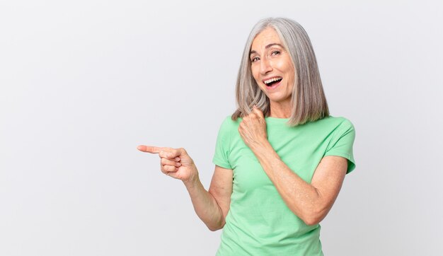 Mujer de cabello blanco de mediana edad que se siente feliz y enfrenta un desafío o celebra y señala hacia un lado