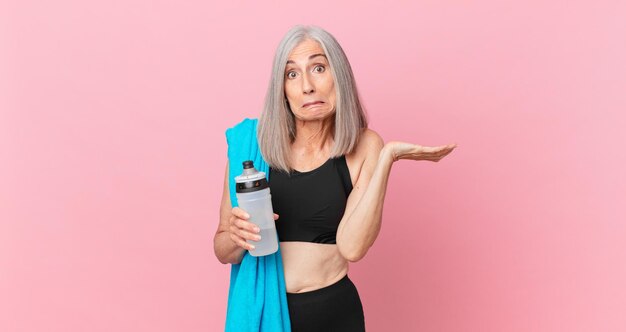 Mujer de cabello blanco de mediana edad que se siente perpleja y confundida y dudando con una toalla y una botella de agua. concepto de fitness