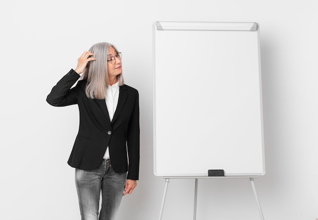 Mujer de cabello blanco de mediana edad que se siente perpleja y confundida, rascándose la cabeza y un espacio de copia de tablero. concepto de negocio