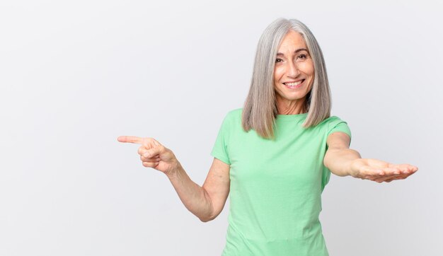 Mujer de cabello blanco de mediana edad sonriendo felizmente con amabilidad y ofreciendo y mostrando un concepto y apuntando hacia un lado