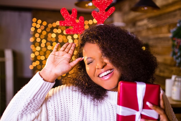 Mujer de cabello castaño de Brasil sostiene una caja de regalo roja cerca de firtree en su apartamento, usa suéter blanco y cuernos de ciervo rojo