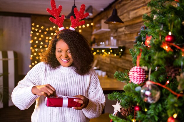 Mujer de cabello castaño de Brasil sostiene una caja de regalo roja cerca de firtree en su apartamento, usa suéter blanco y cuernos de ciervo rojo