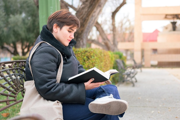 Mujer con cabello corto leyendo un libro sentado en un banco del parque