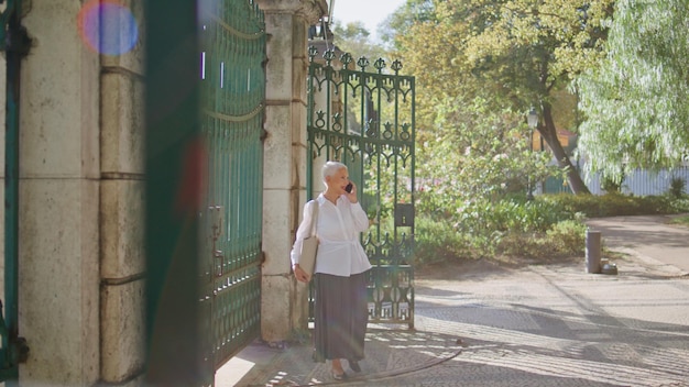 Foto mujer de cabello gris llamando al teléfono móvil esperando a un amigo en la vista del parque a través de la puerta