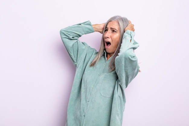 Mujer de cabello gris de mediana edad con la boca abierta, con aspecto horrorizado y conmocionado debido a un terrible error, levantando las manos a la cabeza