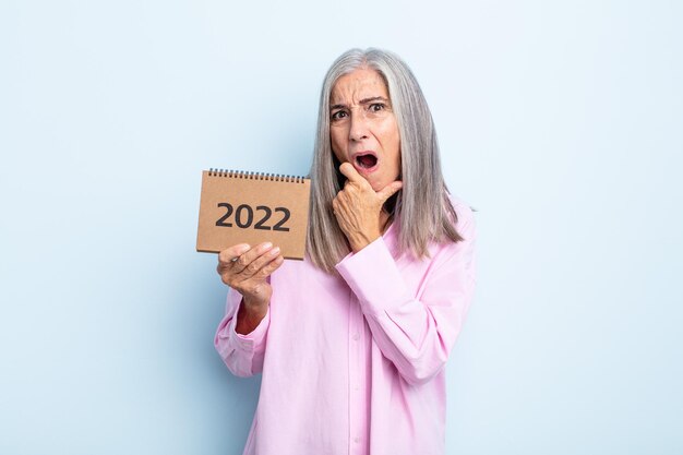 Mujer de cabello gris de mediana edad con la boca y los ojos bien abiertos y la mano en la barbilla concepto de calendario 2022