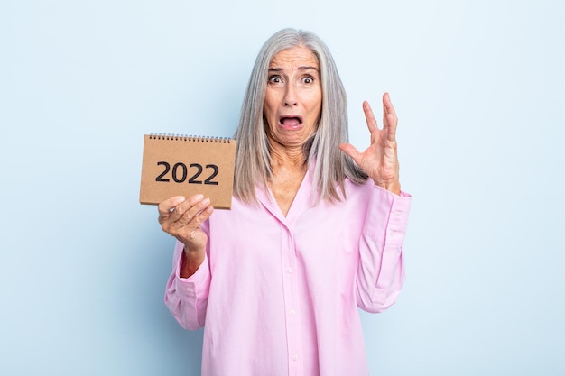 Mujer de cabello gris de mediana edad gritando con las manos en el aire concepto de calendario 2022